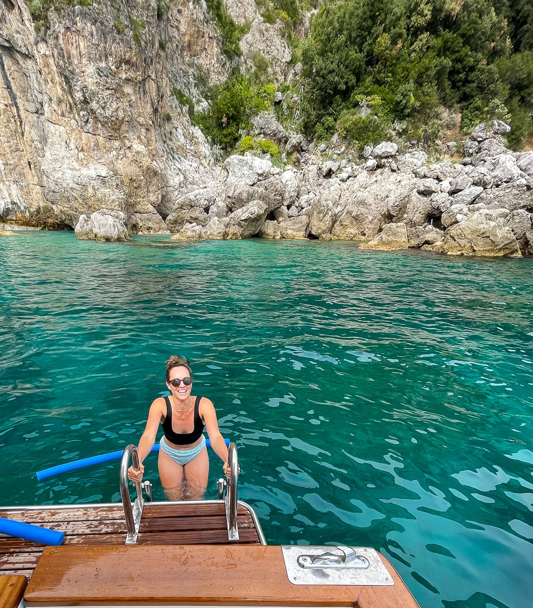 Swimmer climbing a boat ladder from turquoise water by rocky cliffs, a fun thing to do in Procida