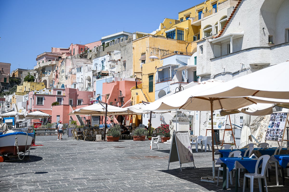 Waterfront restaurants under white umbrellas below colorful houses at Marina Corricella, Procida