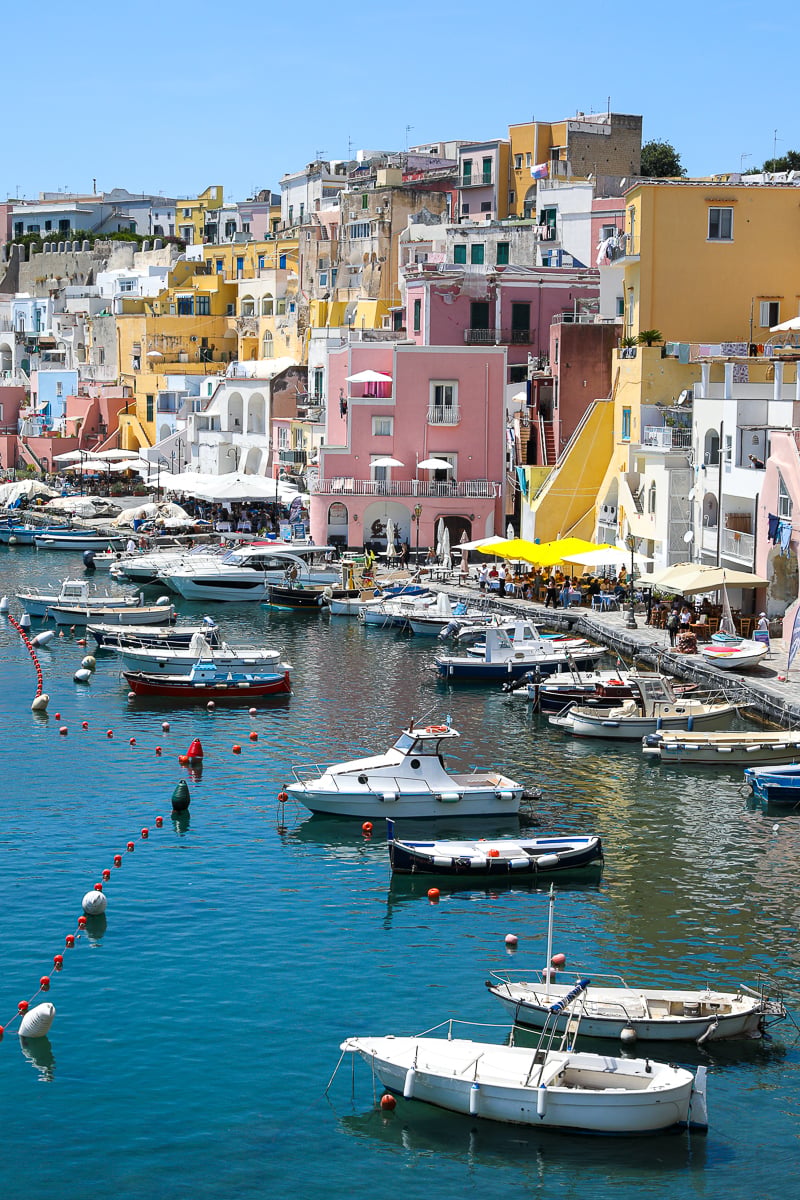 Fishing boats moored in the turquoise harbor below the pastel buildings of Marina Corricella, Procida