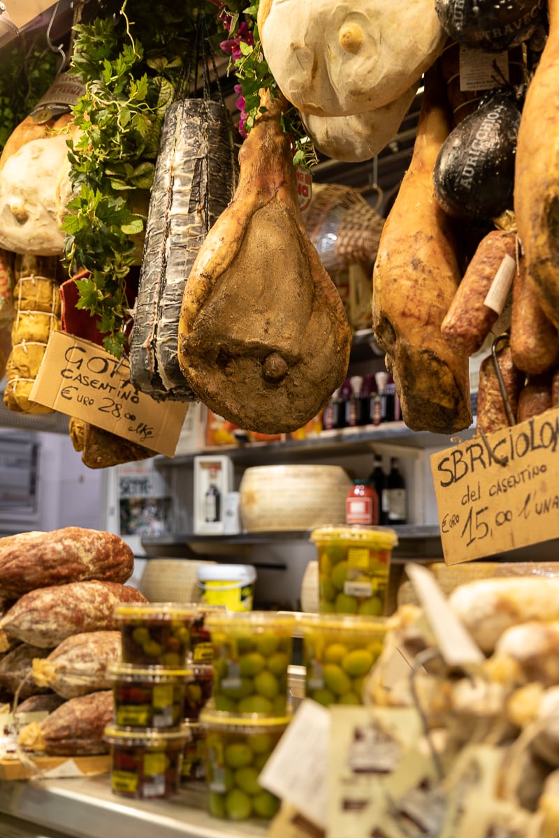 Cured hams, salami, and cheese wheels hanging above a deli counter at Mercato Centrale in Florence.