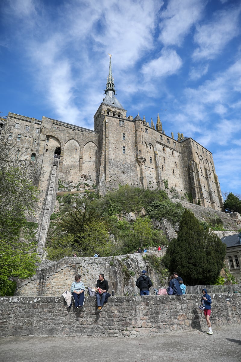 Visitors resting on a stone wall below the ramparts and abbey of Mont Saint-Michel, Normandy