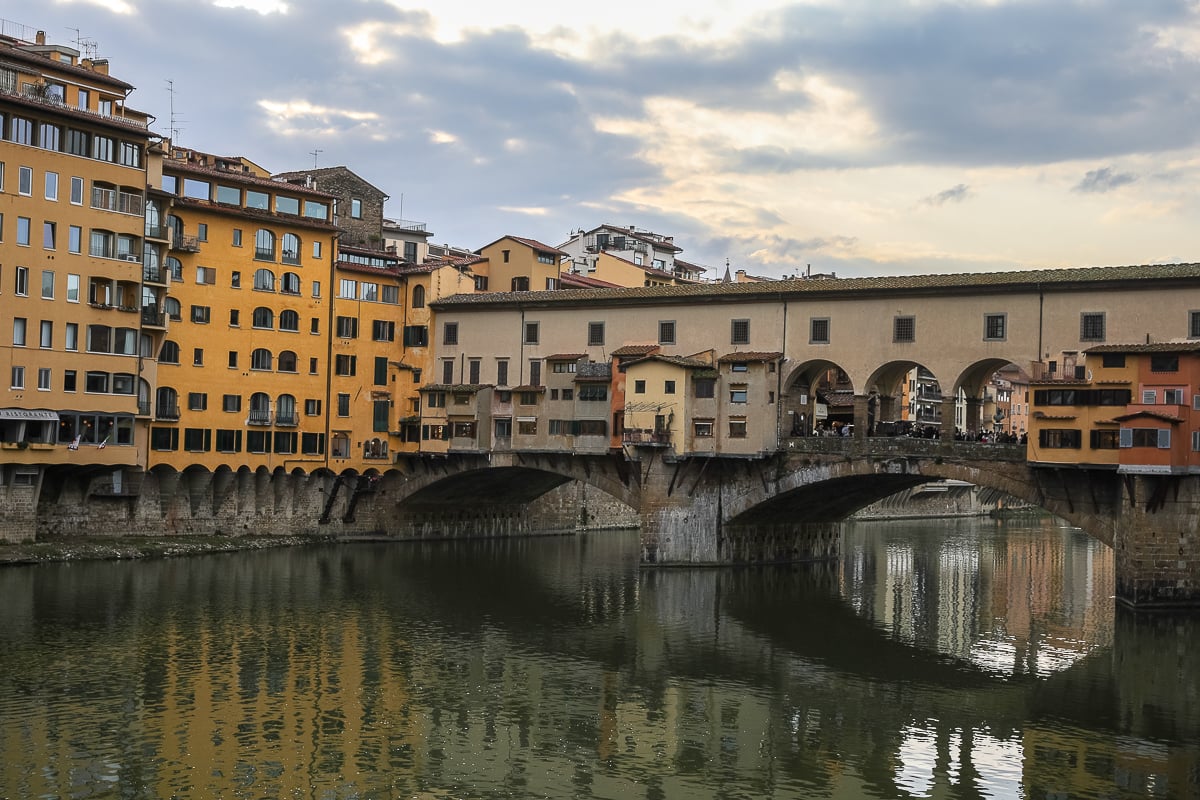 View of the Ponte Vecchio bridge and its shop-lined arches reflected in the Arno River at dusk.