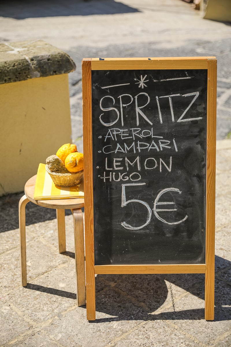 Chalkboard menu for Aperol, Campari and Hugo spritz at 5 euros beside a basket of lemons