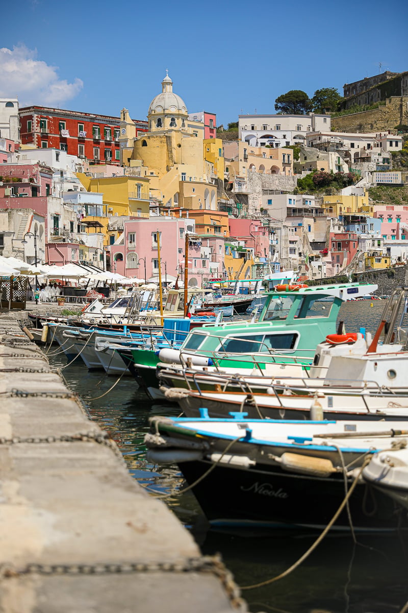 Wooden fishing boats docked along the quay below Corricella's pastel houses and church dome in Procida