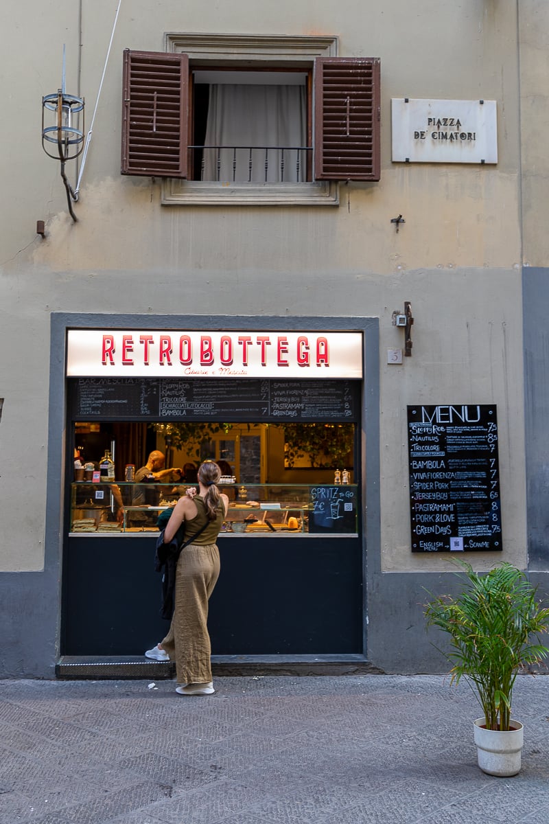 Maddy ordering at the counter of Retrobottega, a small sandwich shop with a red illuminated sign and chalkboard menu.