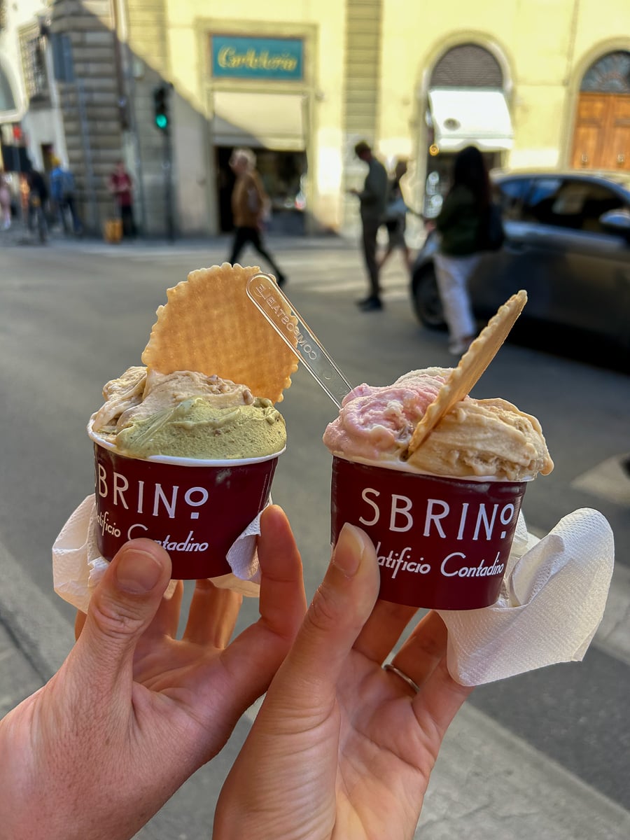 Two hands holding cups of gelato from Sbrino, topped with wafer cookies, on a city street.