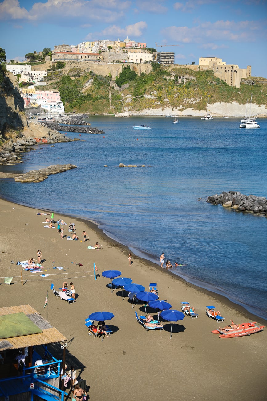 Sunbathers and blue umbrellas on the sand at Spiaggia della Chiaia below Terra Murata in Procida