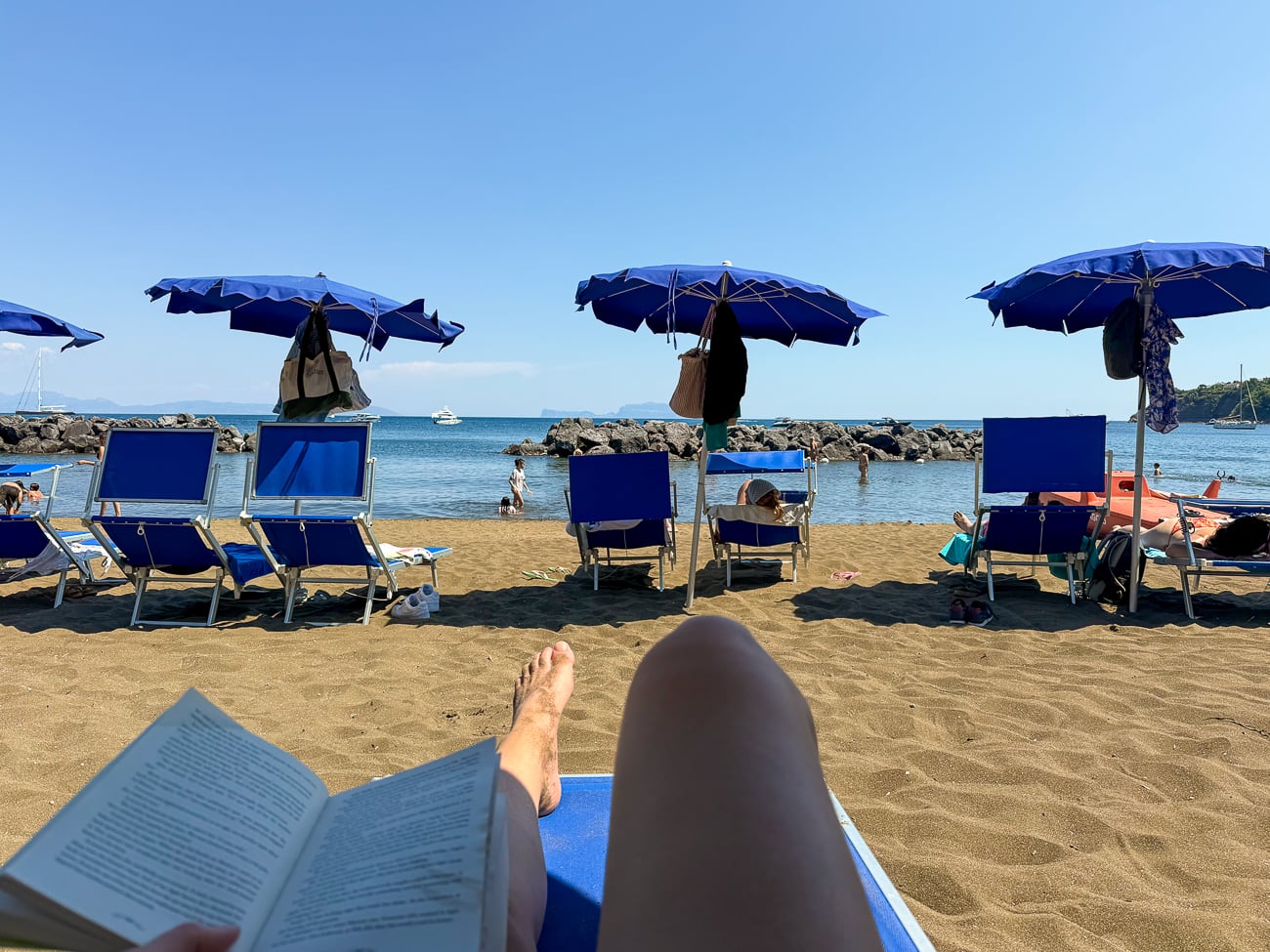 Reading on a sun lounger facing blue umbrellas and the sea at a beach in Procida, a great thing to do