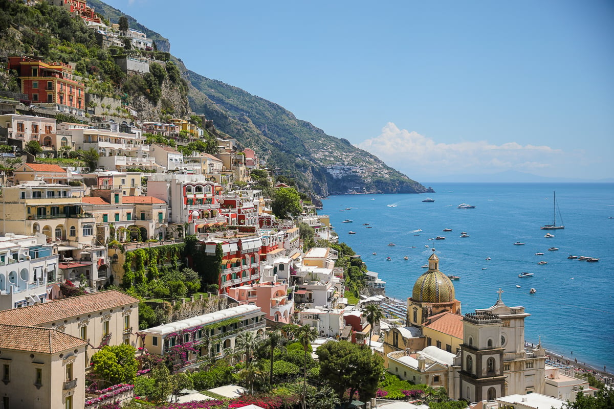Elevated view of Positano's layered pastel buildings, with a majolica-tiled church dome in the foreground and the coast stretching beyond