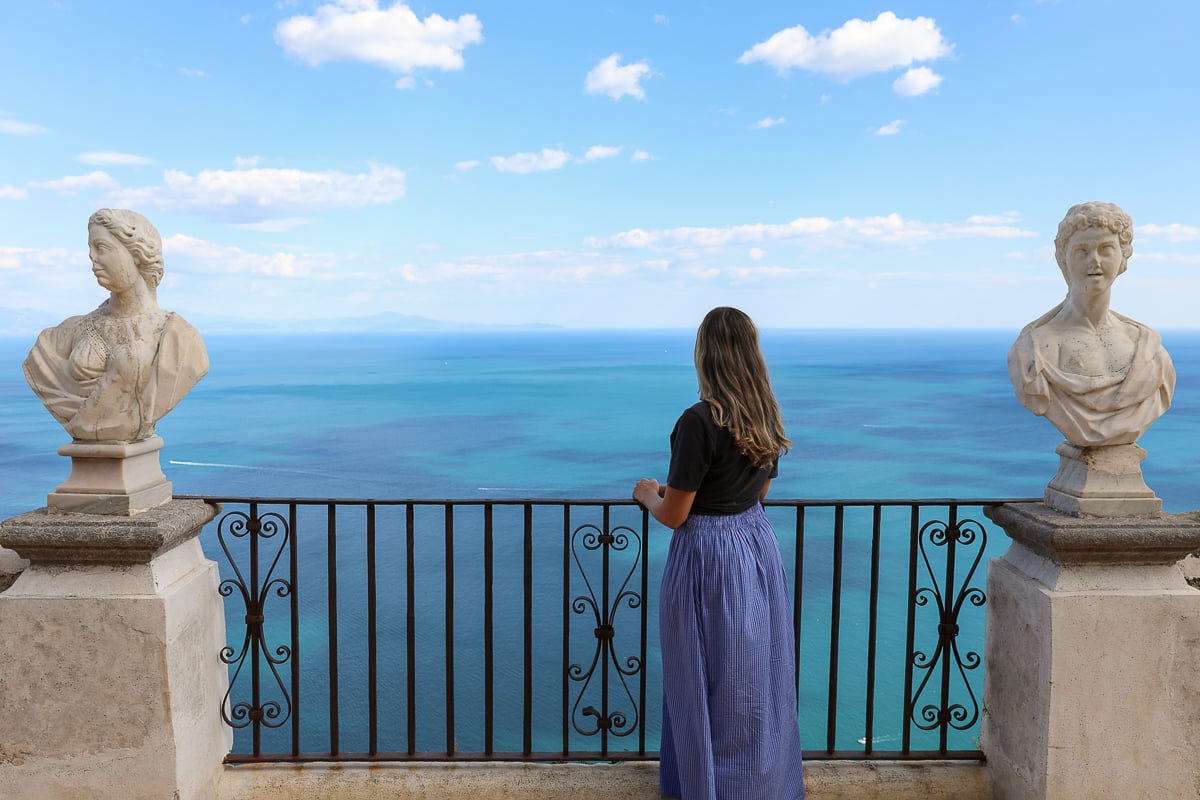 Maddy looking out from a cliffside terrace lined with classical marble busts, overlooking the deep blue Amalfi Coast - during a day trip from Naples