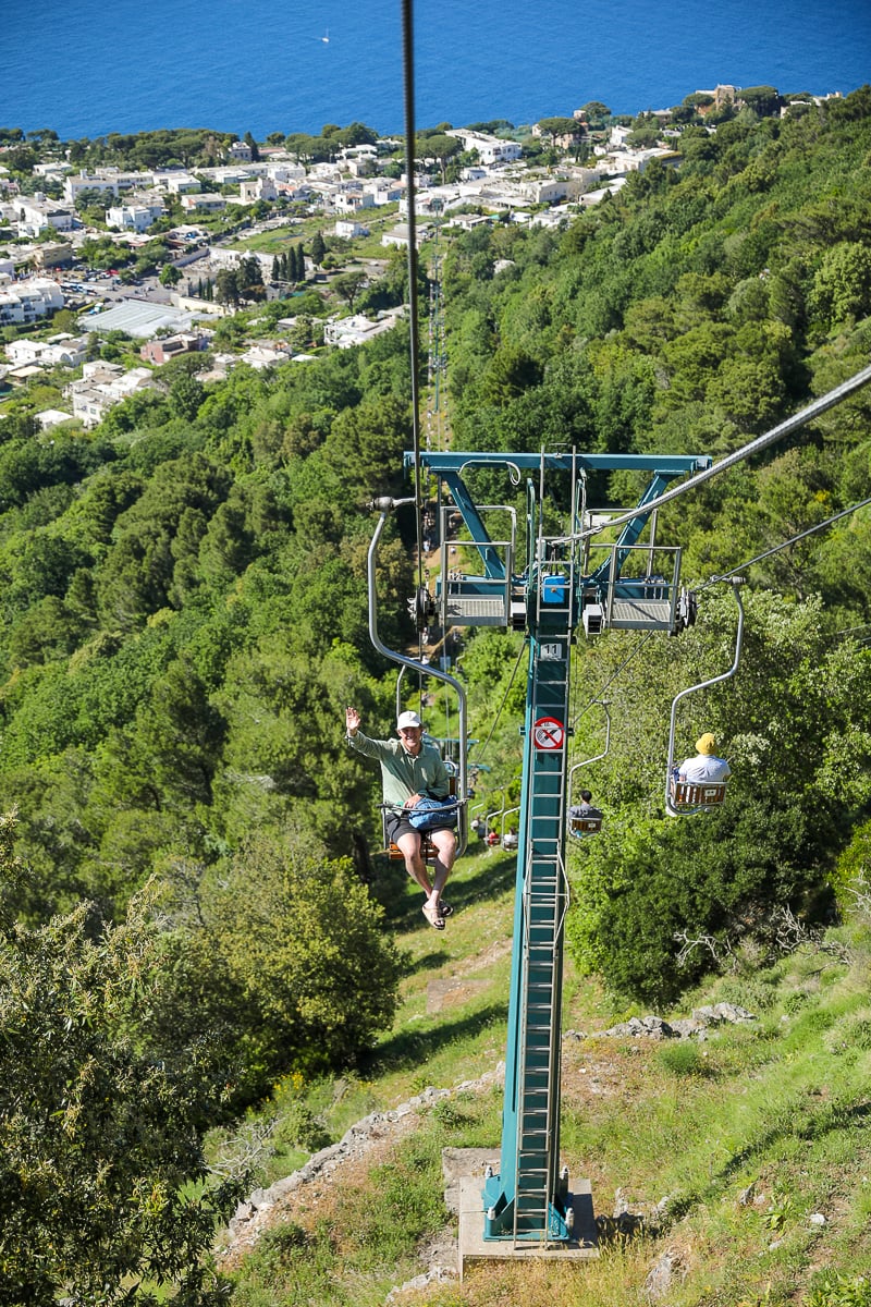 Open-air chairlift carrying passengers up a forested slope on Monte Solaro, with Anacapri's white rooftops visible below