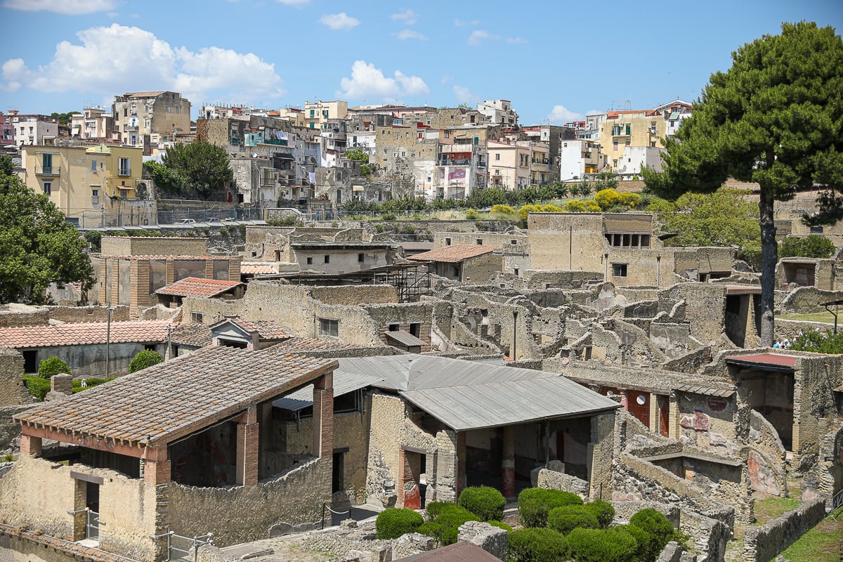 Excavated ruins of ancient Herculaneum, with tiled roofs and stone walls set against a hillside of modern apartments