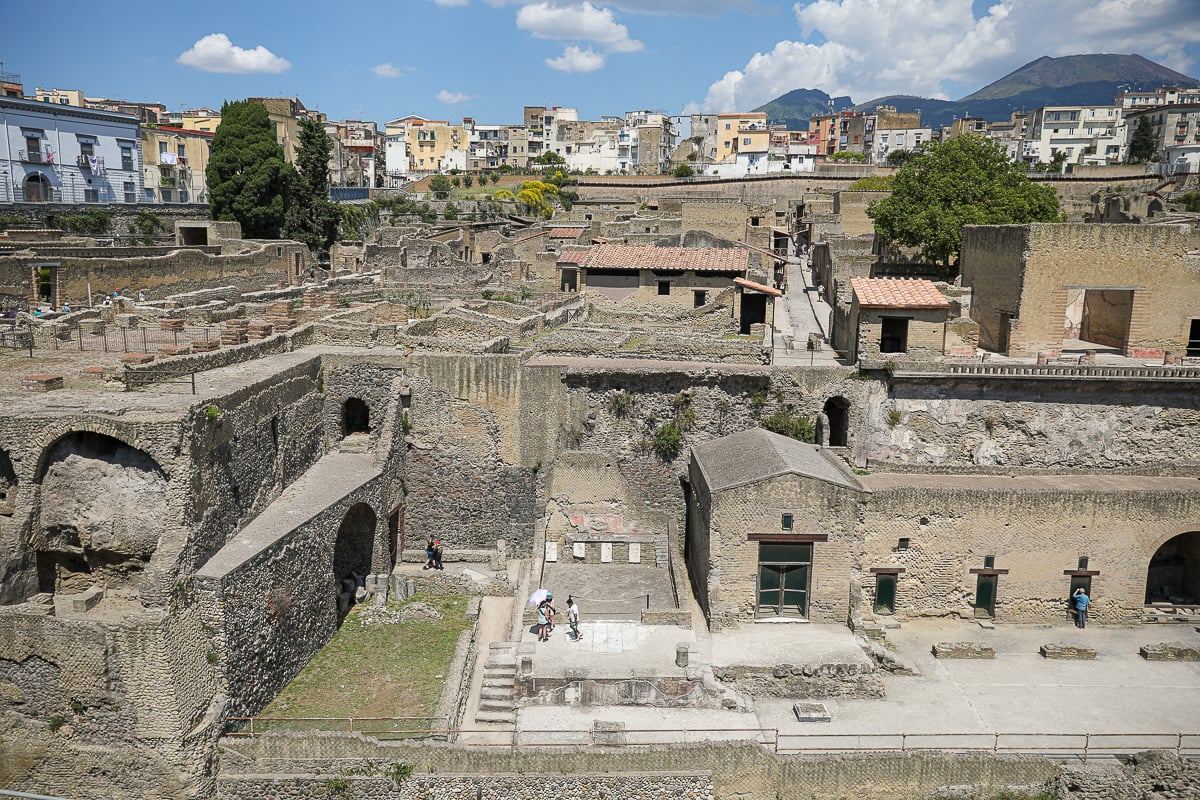 Excavated stone ruins of ancient Herculaneum, with tiled rooftops and arched doorways set against modern apartments and Mt. Vesuvius