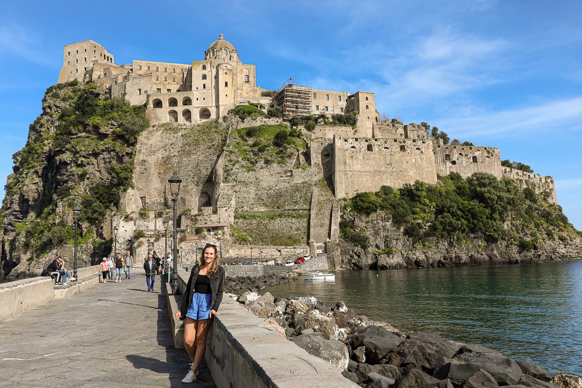 Maddy standing along a stone seawall with the Aragonese Castle's fortified walls and domed rooftop rising behind her