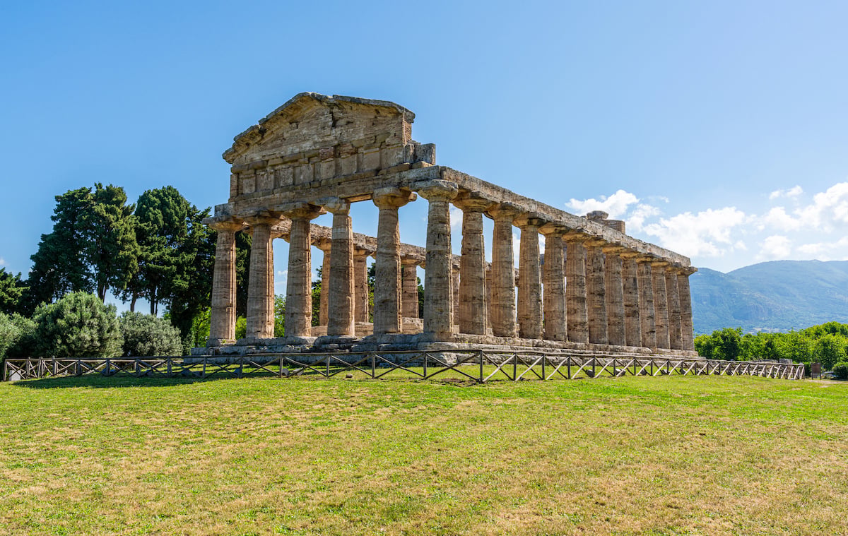 Row of ancient Doric columns at the Temple of Athena in Paestum, set on a grassy field with mountains behind. This is one of the best day trips from Naples by train.