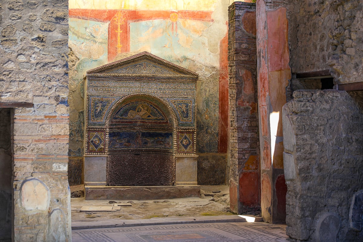 An ornate mosaic-decorated Roman fountain niche inside a Pompeii residence, featuring intricate glass tiles, a pedimented roof, and faded frescoes on the surrounding wall.