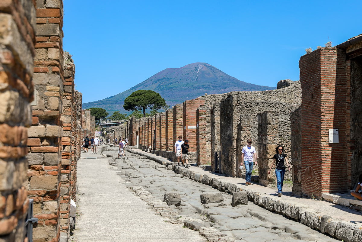 Ancient cobblestone street lined with brick ruins, seen on a classic Pompeii day trip from Naples with Mt. Vesuvius in view