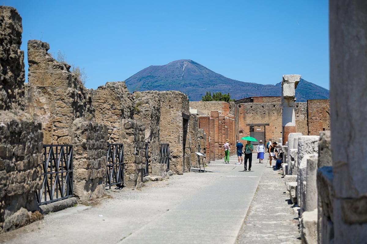 Tourists walking along a stone street through the ruins of Pompeii, with Mt. Vesuvius rising under a clear blue sky
