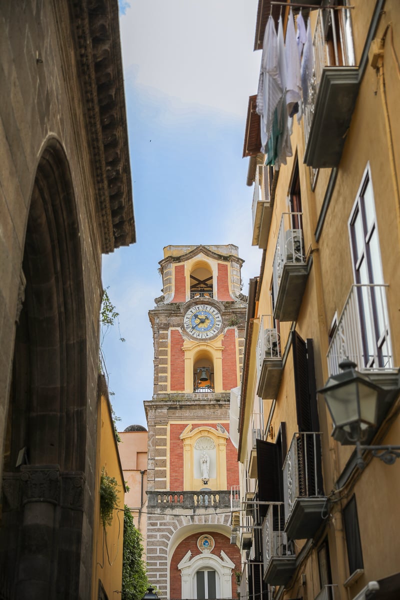 Ornate red-and-yellow Baroque bell tower with a clock face, glimpsed between narrow buildings with laundry hanging from balconies