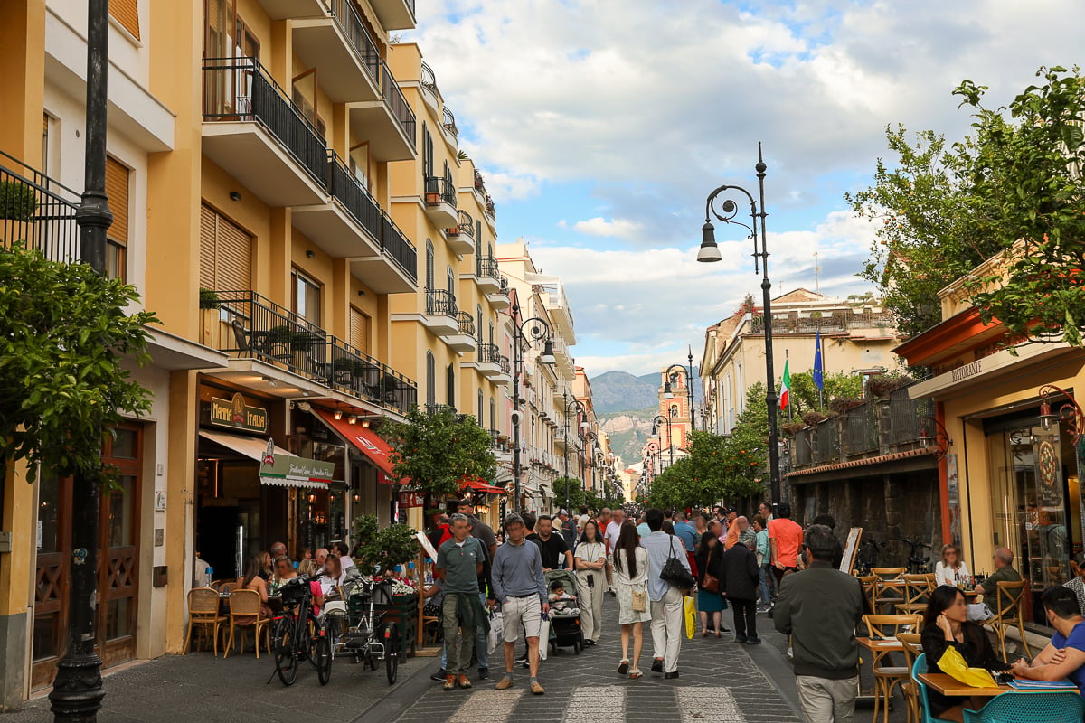 Bustling pedestrian street in Sorrento lined with yellow buildings, outdoor cafes, and shoppers strolling past iron balconies