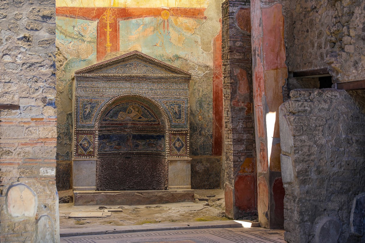 Small marble and mosaic household shrine framed by faded frescoes inside an ancient Pompeii villa
