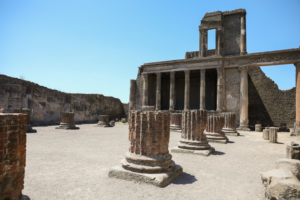 Weathered brick columns and partially restored stone building at the ancient Roman basilica in the Pompeii archaeological site under a clear blue sky.