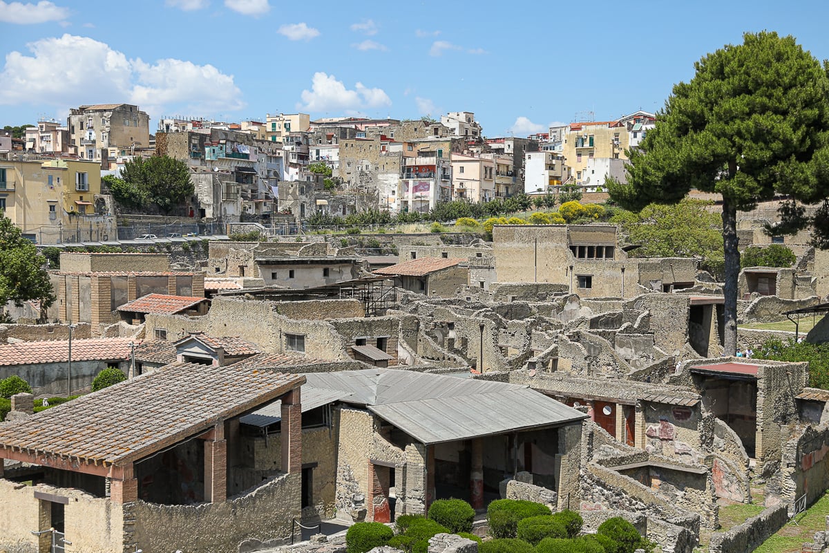Wide-angle view of the excavated ruins of Herculaneum, with preserved stone structures and roof coverings contrasted against the modern Italian town built on the higher ground above.