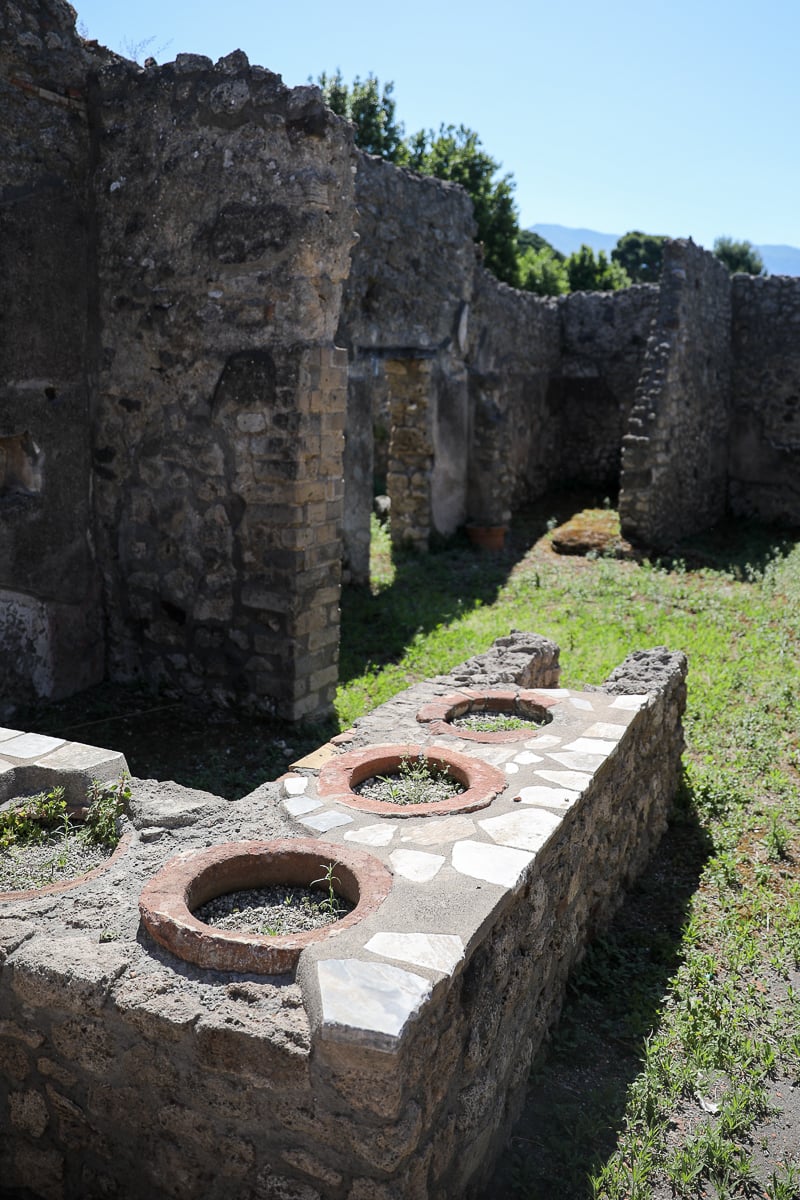 A close-up view of an ancient Roman thermopolium, or snack bar, in Pompeii, featuring stone counters with circular insets that once held clay jars for food.