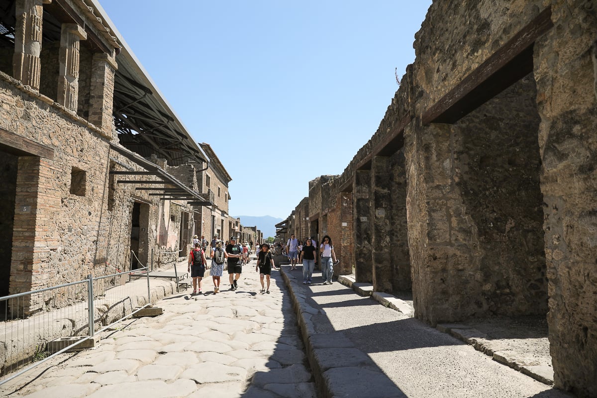 Tourists walking along a cobblestone street in Pompeii, lined with stone ruins and columns under a clear blue sky, with a modern metal roof canopy visible on the left.