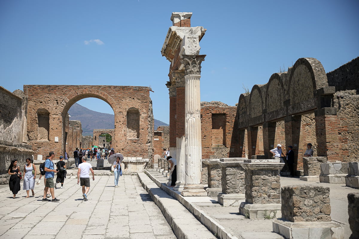 A sunlit street within the Pompeii Forum, showing visitors among red-brick walls and stone columns, with a large stone arch framing distant mountains under a blue sky.