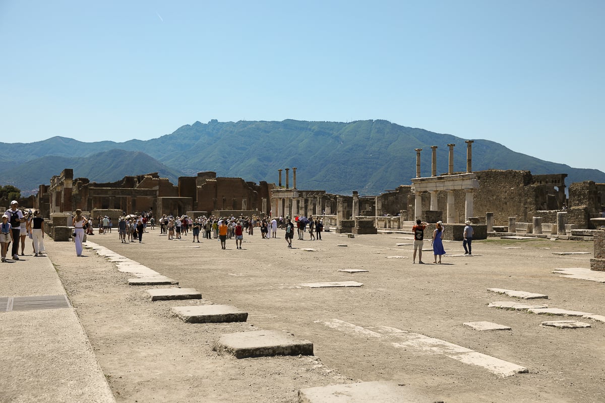 A wide-angle view of the Pompeii Forum, with tourists walking through the dusty plaza among scattered stone ruins and columns, with a large green mountain in the background.