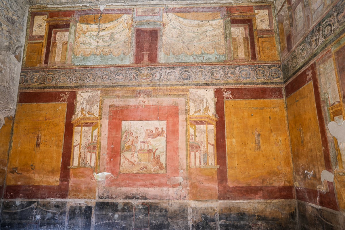 A well-preserved Roman wall painting in the Fourth Style, featuring a central narrative panel surrounded by architectural motifs, rich ochres, deep reds, and geometric borders.