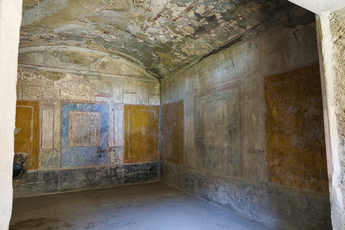 An interior room in a Pompeii villa with well-preserved yellow and blue wall frescoes and a vaulted ceiling showing traces of ancient Roman art.