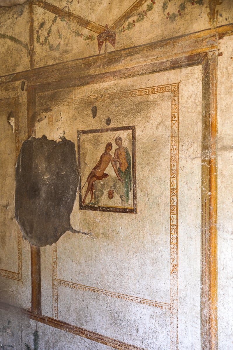 A detailed view of a partially preserved Roman fresco painting on a plaster wall, showcasing a scene with two figures in the style of ancient Pompeian art.