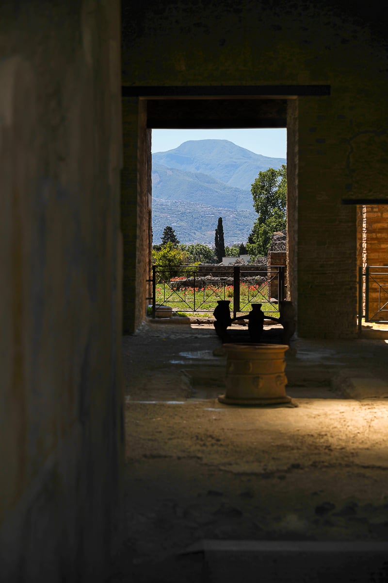 A view through a stone doorway in the Pompeii ruins, with shafts of sunlight illuminating ancient artifacts in the foreground and a distant mountain range under a bright sky.