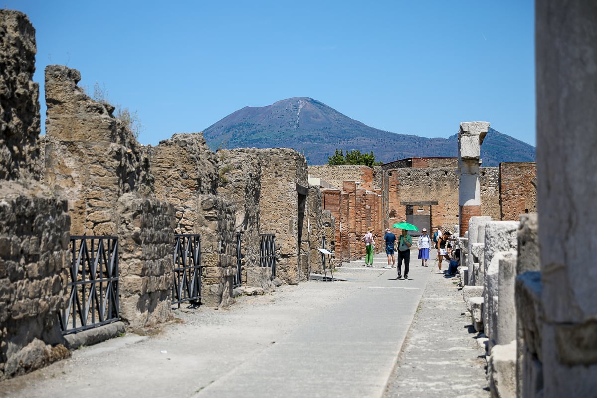 A view of an ancient Roman street in Pompeii, with tourists walking past weathered stone walls and ruins while Mount Vesuvius looms in the distance under a clear blue sky.