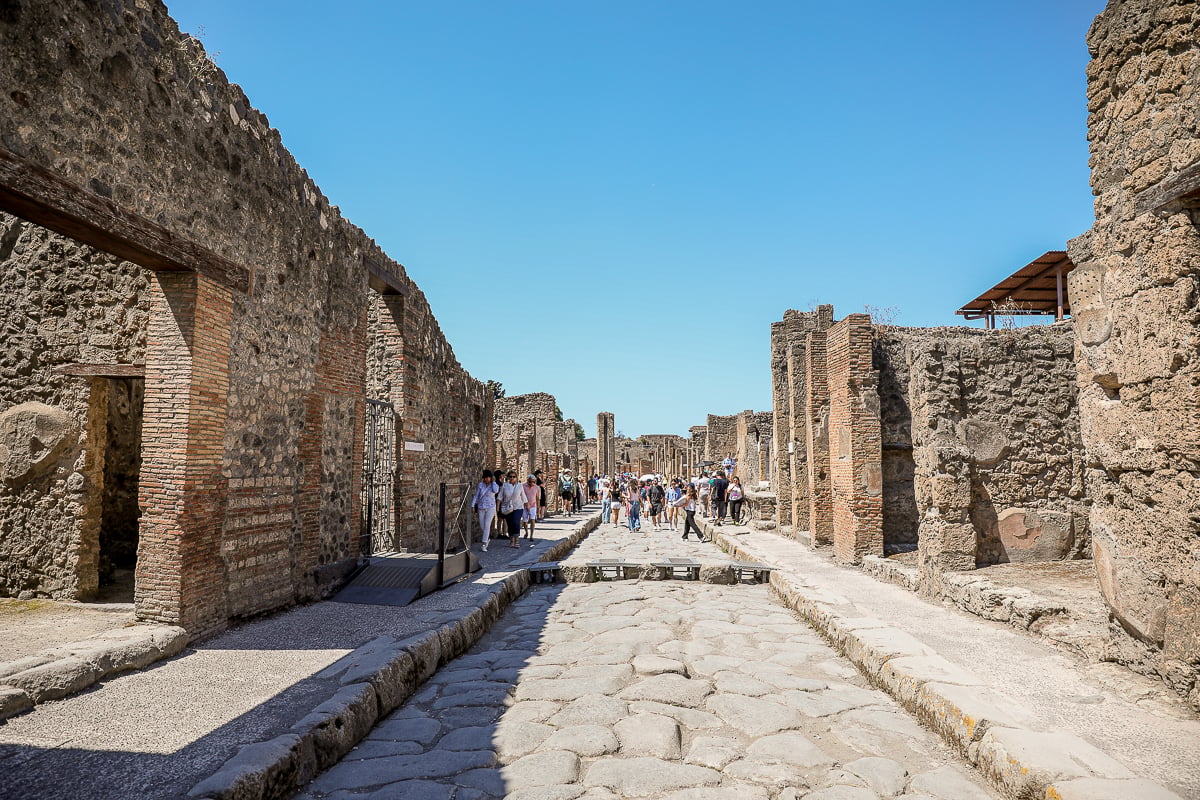 A wide stone-paved street in Pompeii, with visitors exploring the ruins amid weathered masonry walls and remnants of Roman architecture on a sunny day.
