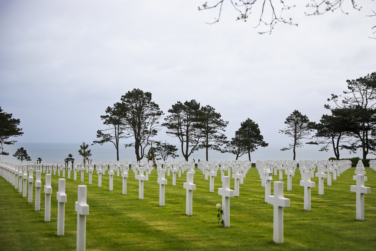Rows of white marble cross headstones standing in perfectly aligned rows on a lush green lawn at the Normandy American Cemetery overlooking the English Channel.