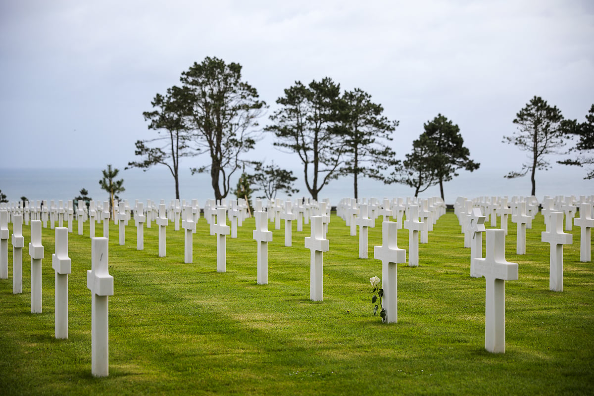 At the Normandy American cememtery, rows of white stone crosses stretch across a green lawn under an overcast sky, with a single white rose at the base of one cross and the English Channel visible in the distance.
