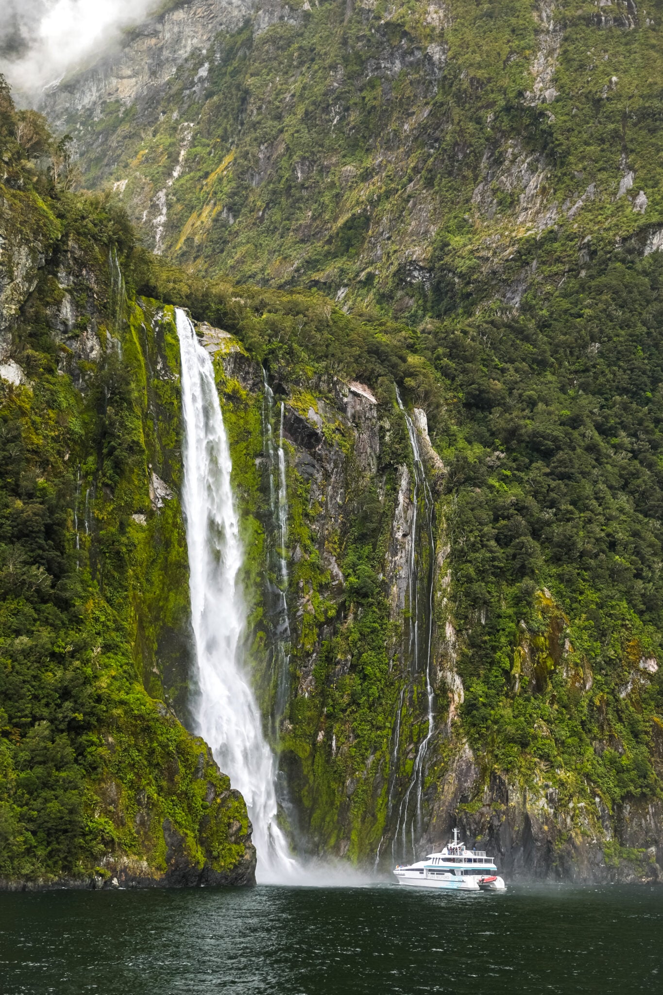 Bowen Falls plunging down a lush green cliff beside a cruise boat in Milford Sound, New Zealand