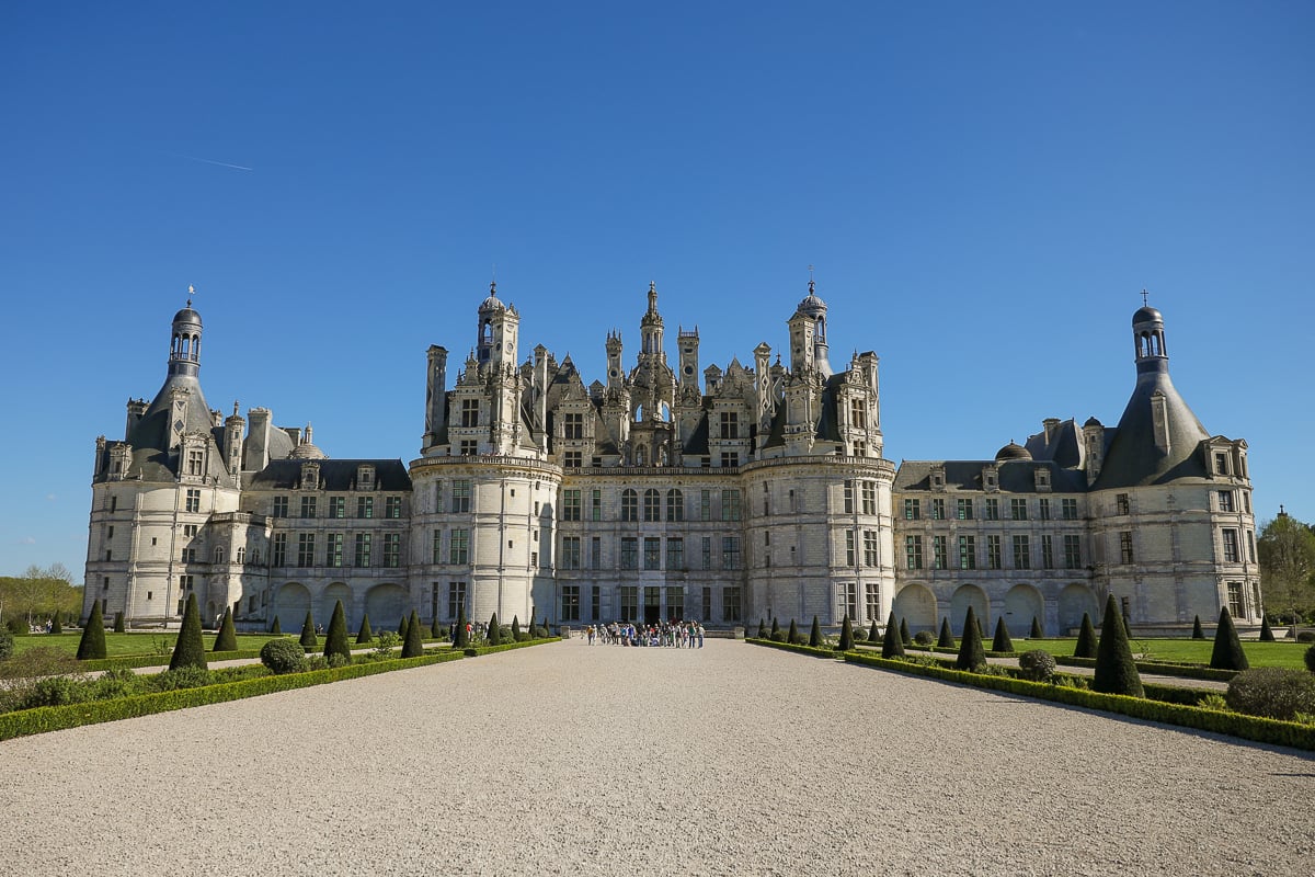 Wide view of Château de Chambord under a clear blue sky, with a gravel path lined by cone-shaped topiaries leading to the castle entrance, where a small group of people stand.