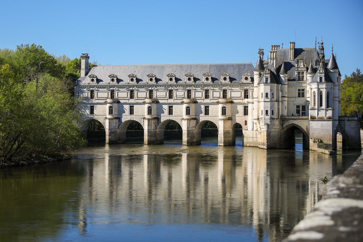 Château de Chenonceau spans the River Cher, its white stone walls, arched supports, and dormer windows reflected in the calm water on a sunny day.