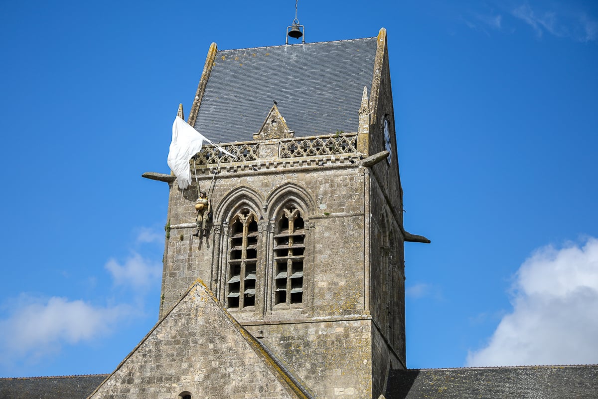 A stone church tower in Sainte-Mère-Église with a replica paratrooper hanging from the roof, commemorating the WWII American airborne drop.