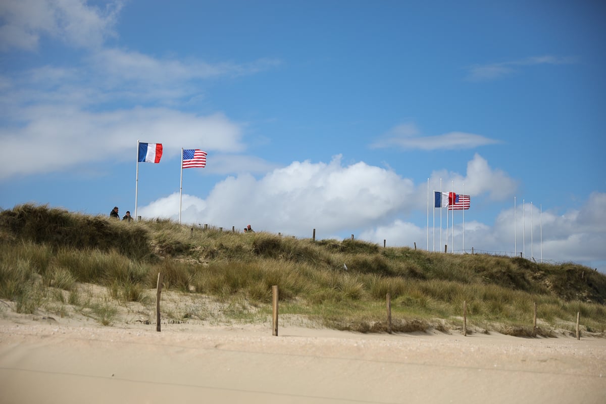 Grassy sand dunes of Utah Beach, Normandy, rise against a blue sky with white clouds, where French and American flags fly from two flagpoles and a few people walk along the ridge.