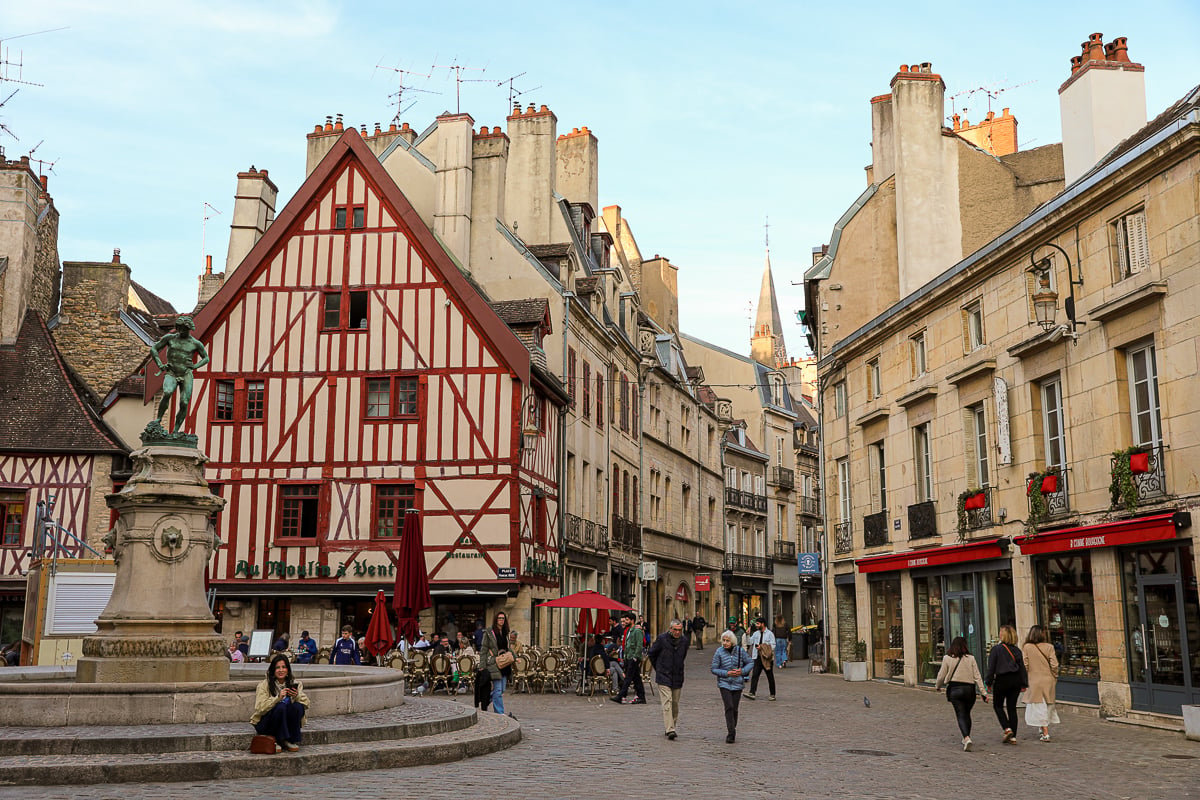 A charming cobblestone square in Dijon, France, where locals and visitors gather at cafe tables near a historic fountain and ornate half-timbered architecture, perfect for those seeking scenic day trips from Paris.