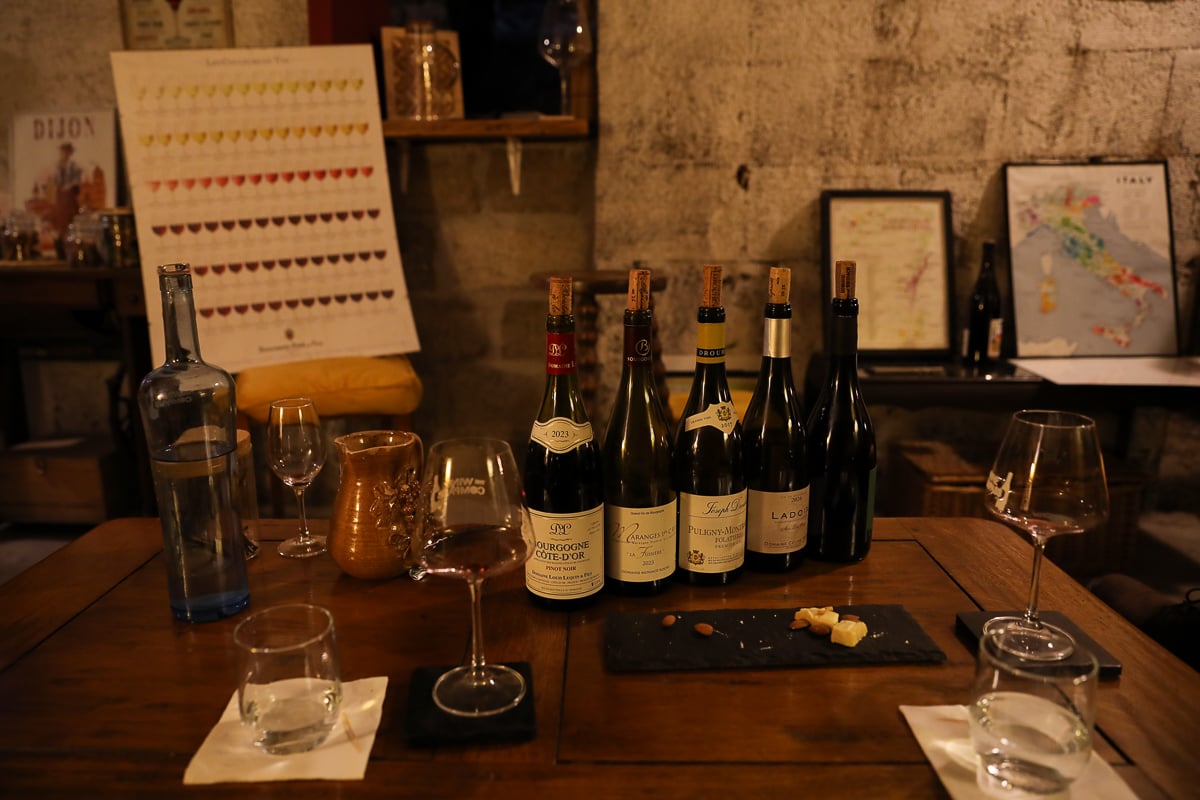 A rustic wooden table set for a wine tasting in Dijon, holding several bottles of red and white wine, glasses, and a small slate platter of cheese and nuts inside a cozy, stone-walled cellar.