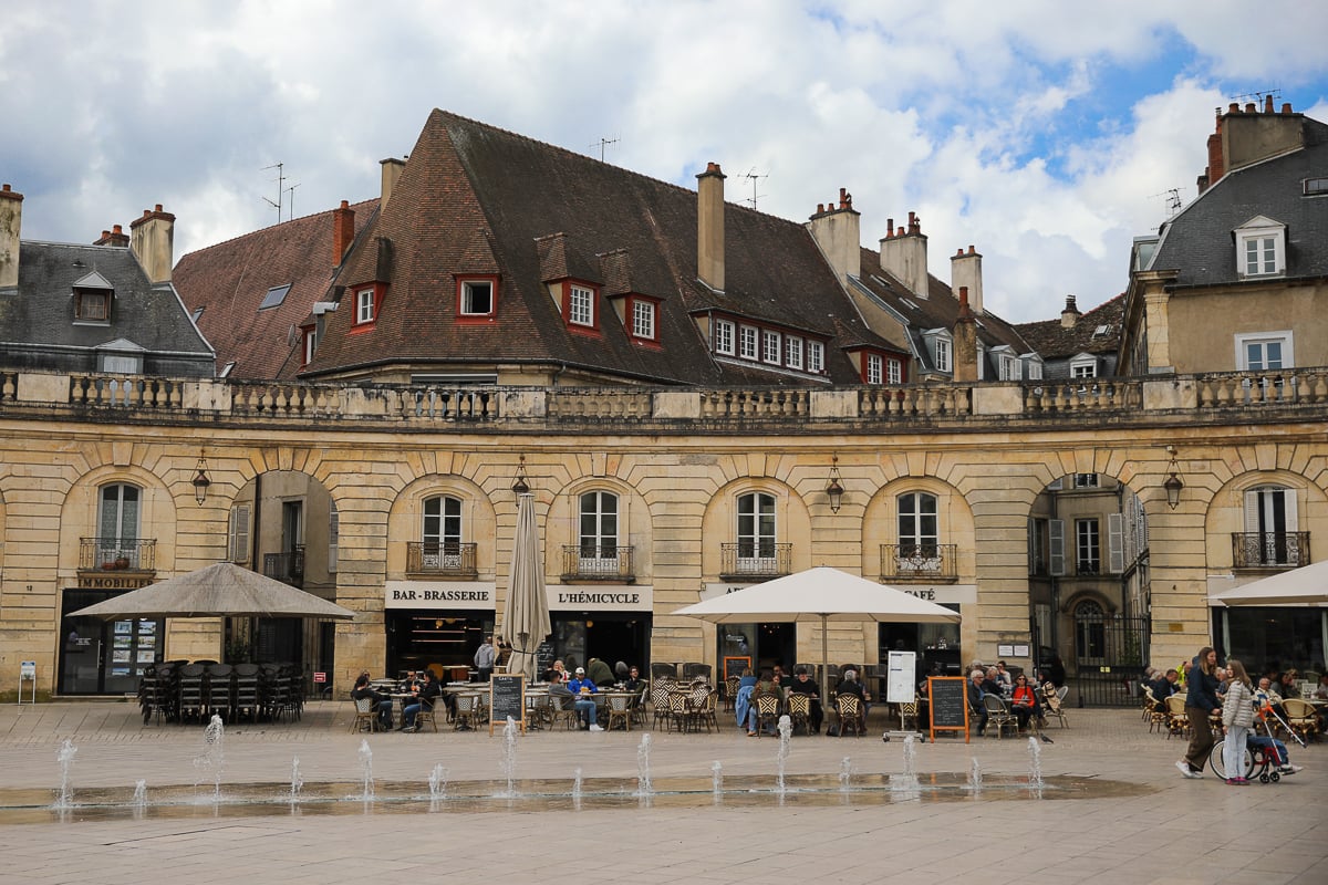 Stone building with arched colonnades housing café terraces around a small fountain in the main square of Dijon, with pedestrians and patrons seated outdoors on a sunny day.