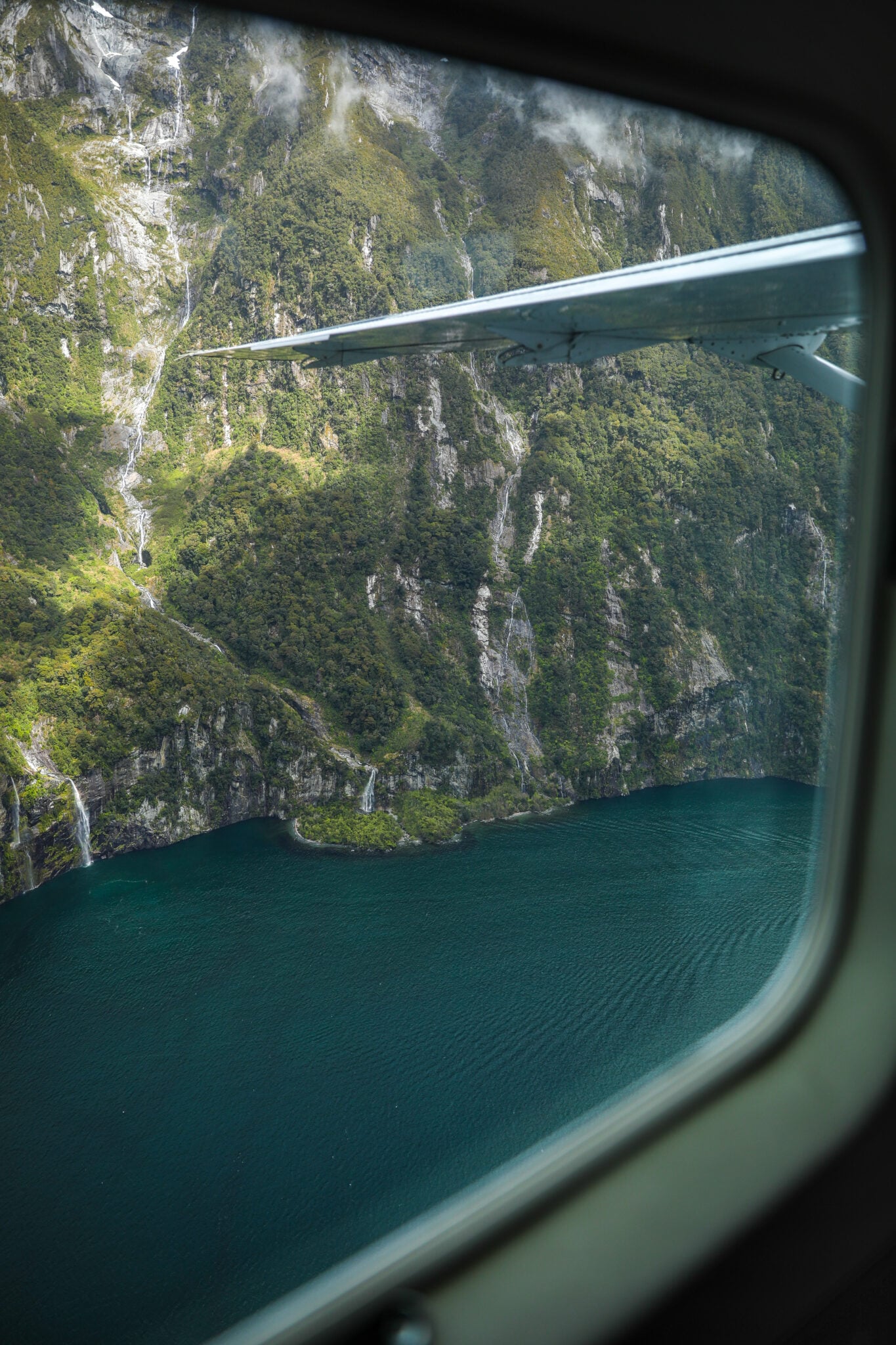 View through a scenic plane window of waterfalls and steep forested cliffs above Milford Sound