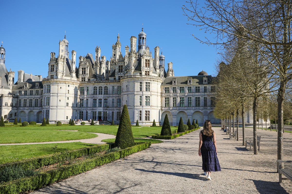 Maddy walking along a gravel path toward Château de Chambord, with manicured lawns and cone-shaped topiaries lining the route under a clear blue sky.