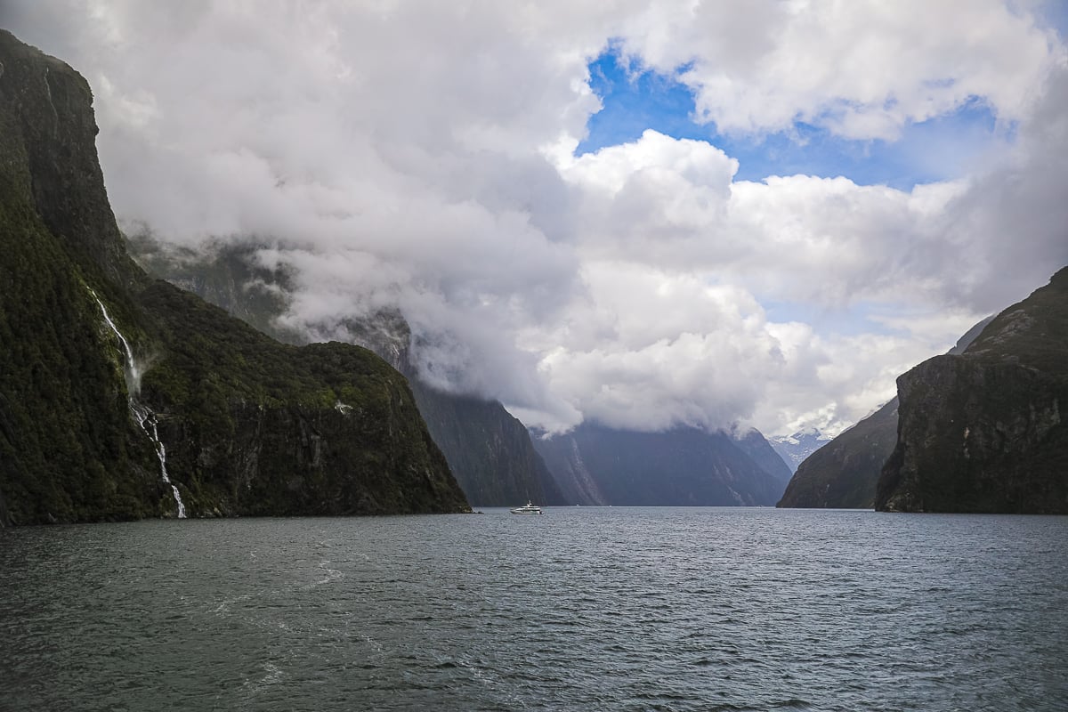 Cruise boat sailing through Milford Sound beneath dramatic clouds, waterfalls and towering mountains